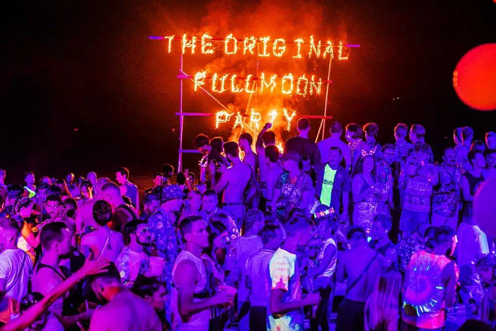 Crowd on the beach at night beneath a burning Original Fullmoon Party sign