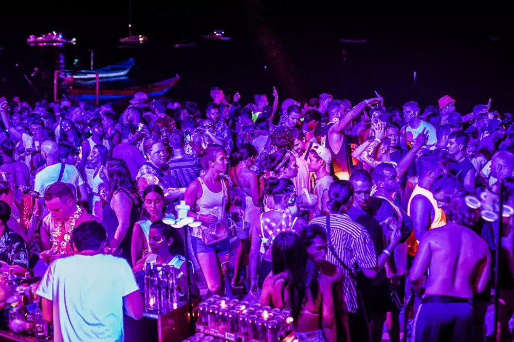 Purple-lit crowd near the shoreline with boats on the water at night
