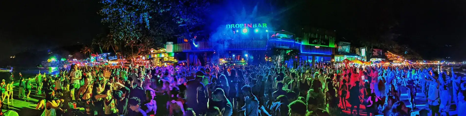 Wide panorama of a huge crowd dancing on the beach in front of neon bars