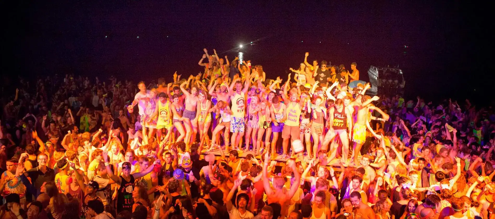 Crowd dancing on a raised platform at a beach party, sea behind at night